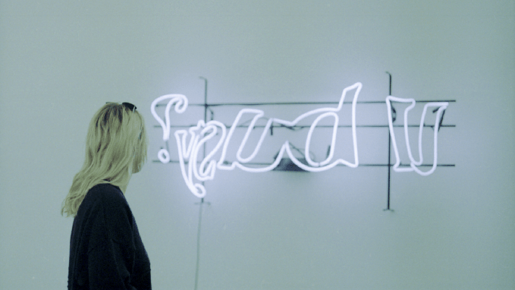 Young woman looking at neon artwork with the words "u busy" in mirrored font