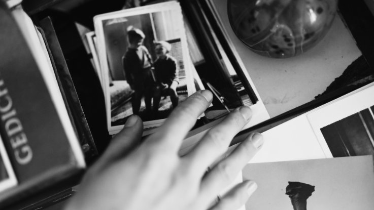 A black-and-white close-up of a hand hovering over a desk with books, pictures and other objects.